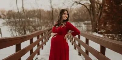 A woman with styled hair and makeup wearing a long red dress standing on a wooden bridge in a snowy landscape.