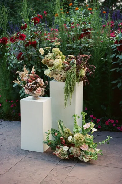 A floral arrangement featuring greenery and flowers displayed on white pedestals in a garden setting.