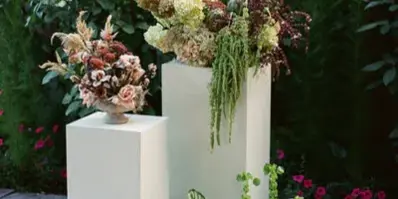 A floral arrangement featuring greenery and flowers displayed on white pedestals in a garden setting.