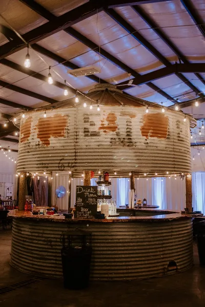 Interior view of a rustic wedding venue bar made from a corrugated metal silo with string lights and a drink menu.