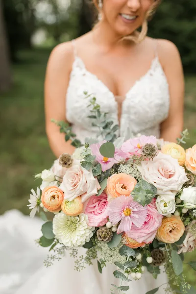 A close-up of a bride holding a large, colorful wedding bouquet featuring pink and peach roses, ranunculus, and eucalyptus.