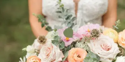 A close-up of a bride holding a large, colorful wedding bouquet featuring pink and peach roses, ranunculus, and eucalyptus.