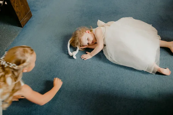A young girl in a white tulle dress lies on a blue carpet while another child looks at her, captured in a candid, documentary style.