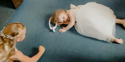A young girl in a white tulle dress lies on a blue carpet while another child looks at her, captured in a candid, documentary style.