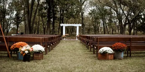 An outdoor wedding ceremony setup featuring a white arch, rows of wooden benches, and floral arrangements in a wooded area.