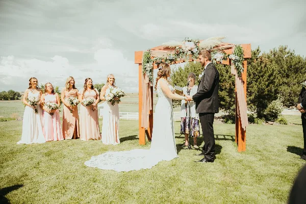 A bride and groom exchanging vows under a floral arch on a grassy lawn with bridesmaids standing to the side.