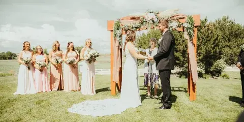A bride and groom exchanging vows under a floral arch on a grassy lawn with bridesmaids standing to the side.