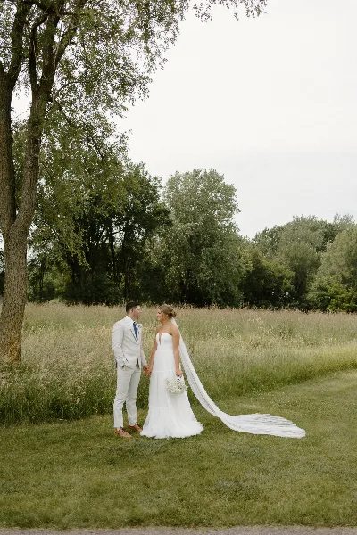 A bride and groom standing in a grassy field holding hands, with the bride wearing a white wedding dress and veil.