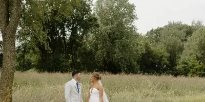 A bride and groom standing in a grassy field holding hands, with the bride wearing a white wedding dress and veil.