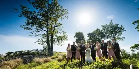 A wedding party consisting of a bride, groom, and attendants posing outdoors on a grassy hill under a bright blue sky.