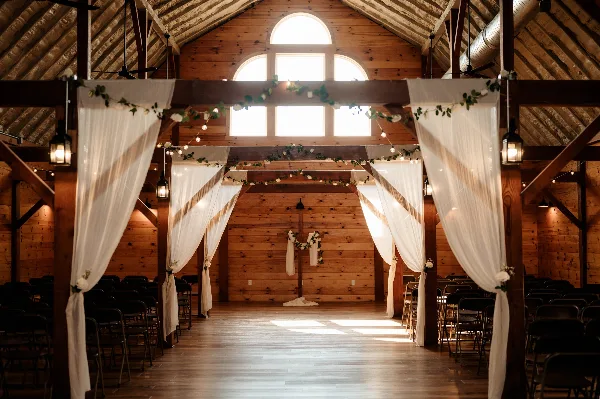 Interior of a rustic wooden barn event center set up for a wedding ceremony with white drapery and floral decorations.