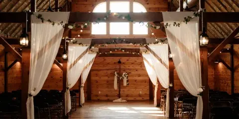 Interior of a rustic wooden barn event center set up for a wedding ceremony with white drapery and floral decorations.