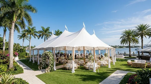 A large white event tent with multiple peaks set up on a grassy lawn near a body of water, containing tables and chairs for a gathering.
