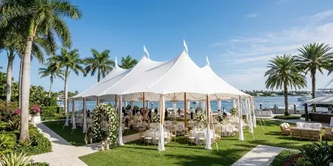 A large white event tent with multiple peaks set up on a grassy lawn near a body of water, containing tables and chairs for a gathering.