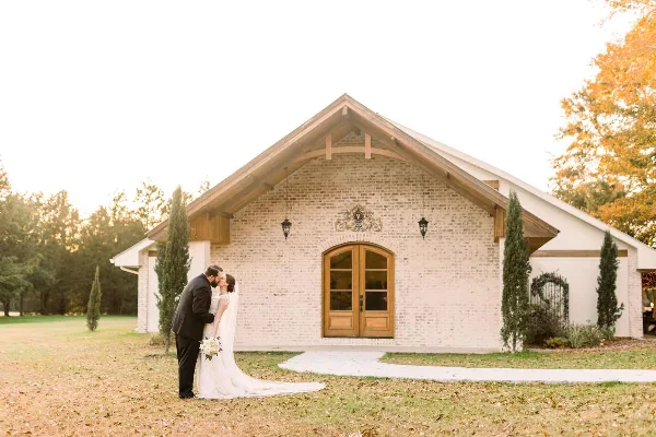 A bride and groom kissing in front of a whitewashed brick wedding venue with wooden beams and arched doors.