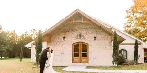 A bride and groom kissing in front of a whitewashed brick wedding venue with wooden beams and arched doors.