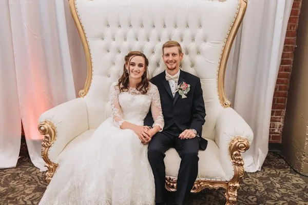 A bride and groom sitting together on a large white and gold throne chair in a wedding venue.