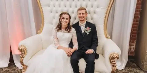 A bride and groom sitting together on a large white and gold throne chair in a wedding venue.