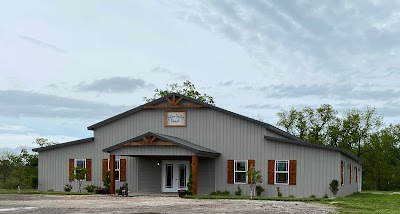 Exterior view of a large, grey metal event barn with a covered entrance and red shutters, set in a rural landscape.