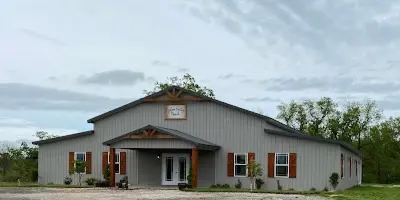 Exterior view of a large, grey metal event barn with a covered entrance and red shutters, set in a rural landscape.