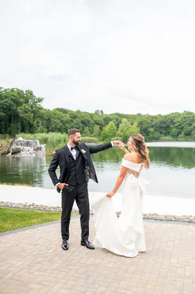 A bride and groom standing on a paved patio near a lake, with the groom twirling the bride by the hand.