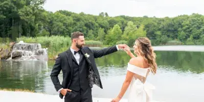 A bride and groom standing on a paved patio near a lake, with the groom twirling the bride by the hand.