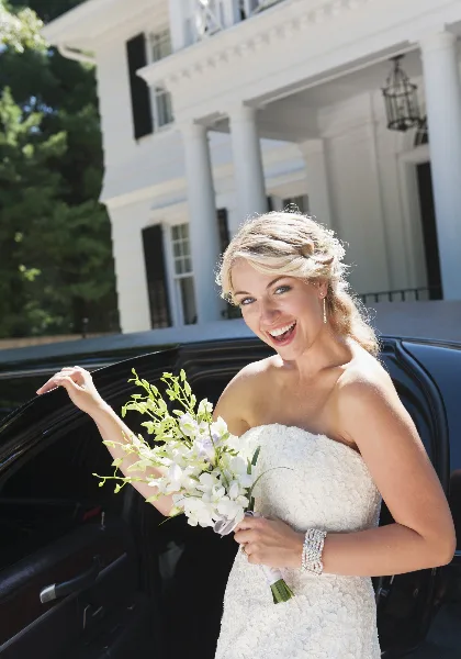 A smiling bride in a white wedding dress holding a bouquet of white flowers, standing next to a black limousine.