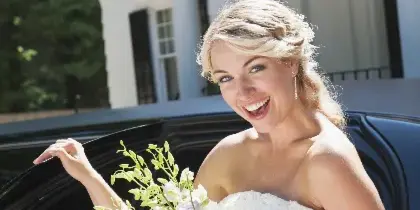 A smiling bride in a white wedding dress holding a bouquet of white flowers, standing next to a black limousine.