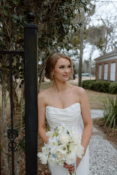 A bride in a white strapless gown holding a white floral bouquet, standing outdoors near a black iron gate.