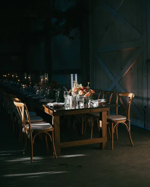 A long wooden dining table set for a wedding reception in a barn, featuring floral arrangements, candles, and wooden chairs.