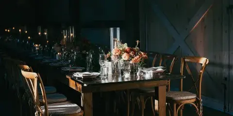 A long wooden dining table set for a wedding reception in a barn, featuring floral arrangements, candles, and wooden chairs.