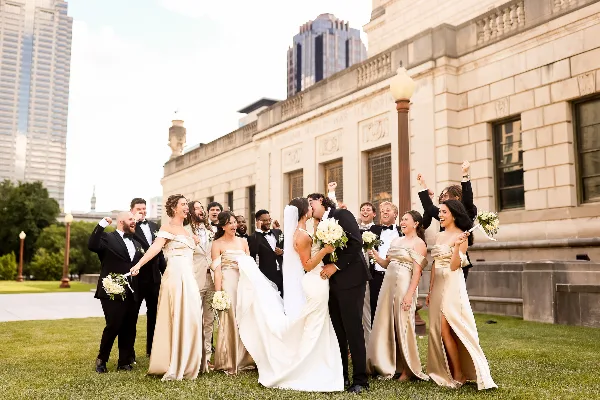 A bride and groom share a kiss in front of a stone building while their wedding party celebrates around them.