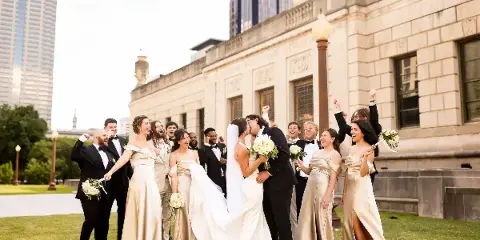 A bride and groom share a kiss in front of a stone building while their wedding party celebrates around them.