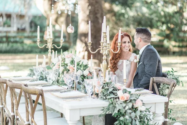 A wedding table setting with floral centerpieces, tall candles, and glassware, with a bride and groom seated in the background.