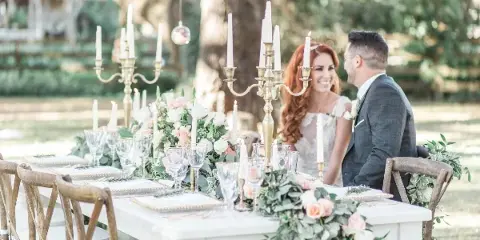 A wedding table setting with floral centerpieces, tall candles, and glassware, with a bride and groom seated in the background.