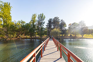 A wooden pedestrian bridge with red railings crossing a calm body of water on a sunny day, surrounded by green trees.