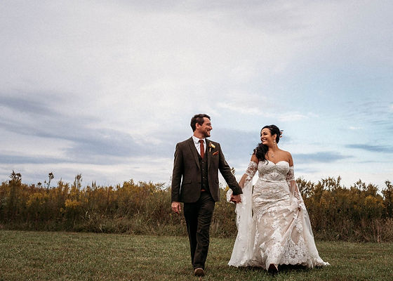 A bride and groom walking hand-in-hand in a grassy field with tall grasses in the background.