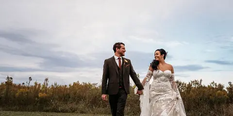 A bride and groom walking hand-in-hand in a grassy field with tall grasses in the background.
