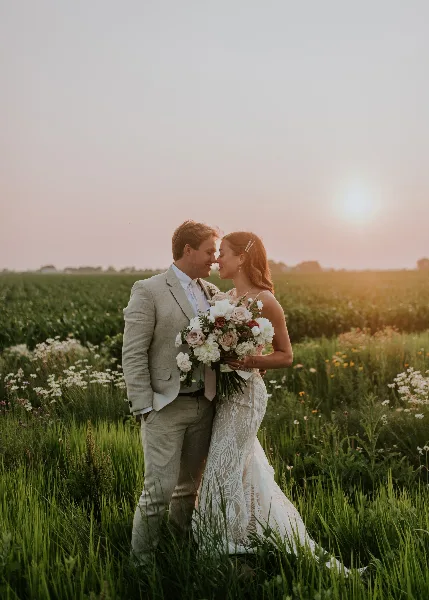 A bride and groom standing in a field of tall grass and wildflowers at sunset, with the bride holding a bouquet.