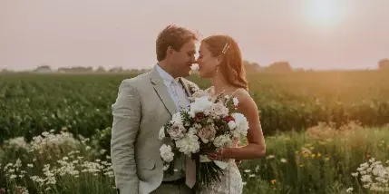 A bride and groom standing in a field of tall grass and wildflowers at sunset, with the bride holding a bouquet.