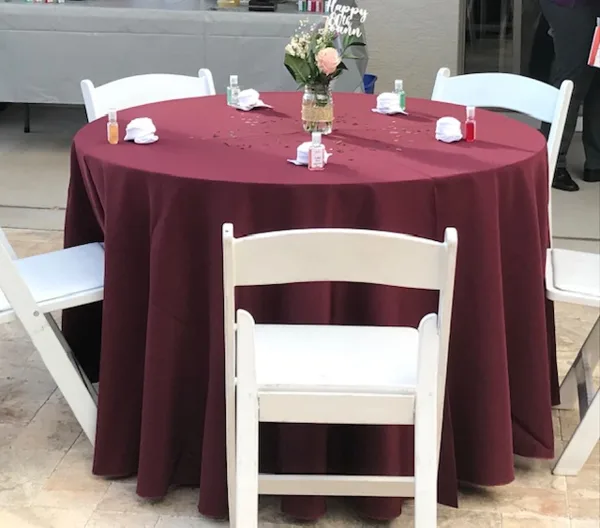 A round event table with a maroon tablecloth, white chairs, and a floral centerpiece.