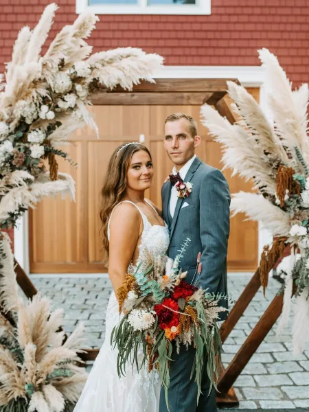 A bride and groom posing in front of a rustic wooden wedding arch decorated with pampas grass and seasonal flowers.