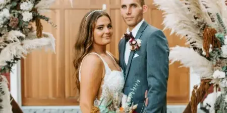 A bride and groom posing in front of a rustic wooden wedding arch decorated with pampas grass and seasonal flowers.