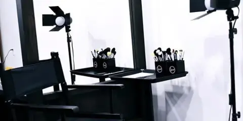 A professional makeup station featuring a director's chair, a large mirror, professional lighting, and makeup brushes.