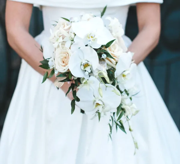A close-up of a bride in a white gown holding a bouquet of white orchids and cream roses.