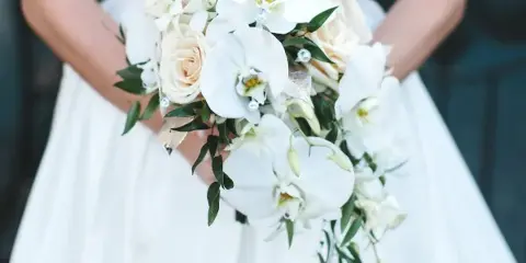 A close-up of a bride in a white gown holding a bouquet of white orchids and cream roses.