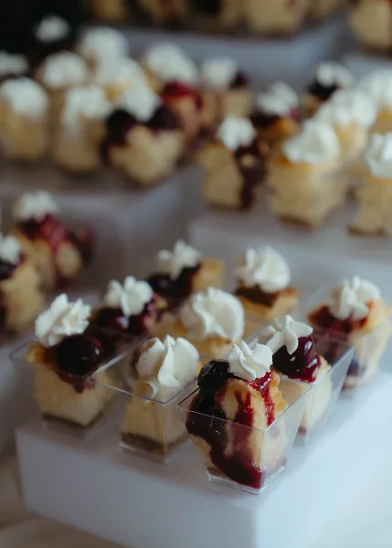 Rows of small dessert bites topped with whipped cream and red fruit glaze, served in clear plastic containers on a white stand.
