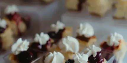 Rows of small dessert bites topped with whipped cream and red fruit glaze, served in clear plastic containers on a white stand.