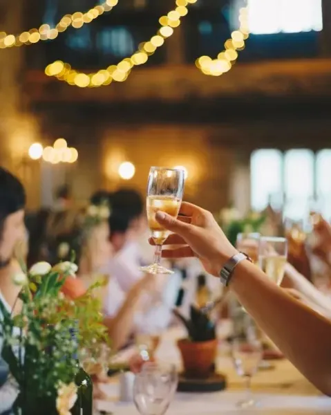 A hand holding a glass of champagne in a toast at a wedding reception with string lights in the background.