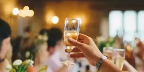 A hand holding a glass of champagne in a toast at a wedding reception with string lights in the background.